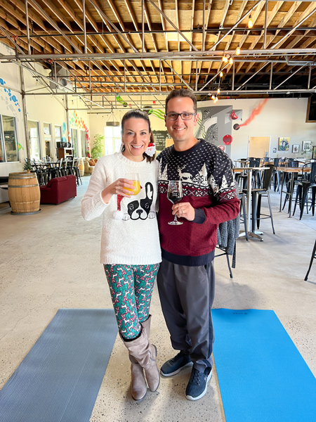 Christine and Tom, a white brunette female in a dog Christmas sweater with reindeer leggings and a white brunette male with glasses in ugly holiday sweater, standing with red wine and a mimosa next to yoga mats at pleb urban winery in Asheville, NC