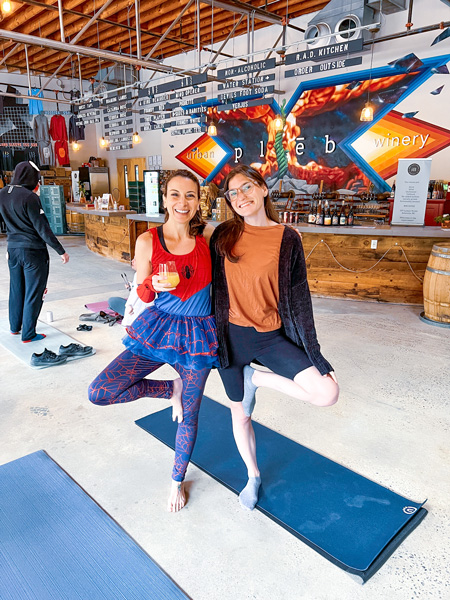 Christine and Sarah, two white, brunette women in yoga clothes - and one with Halloween Spiderman themed outfit - in front of yoga mats with mimosa; bar with murals blurred in the background at pleb urban winery in Asheville's River Arts District