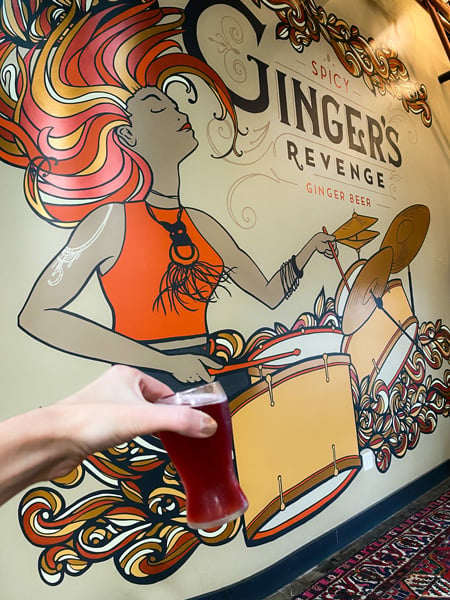 Gingers Revenge Asheville NC with white hand holding up flight glass with red ginger beer in front of logo and mural with red, yellow and orange haired woman playing the drums