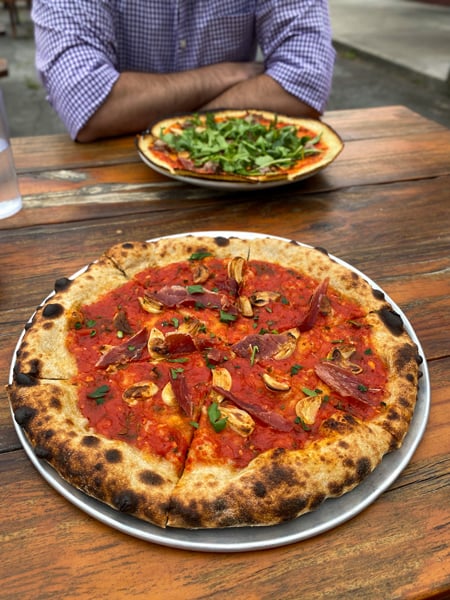 All Souls Pizza Asheville NC with cheeseless pizza with red sauce, meat, basil, and roasted garlic on picnic table with gluten-free polenta crust pizza in background with man in white and purple striped shirt