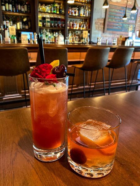 Bone And Broth Asheville Cocktails with one low ball and one high glass of pink and orange cocktails on table with bar blurred in the background