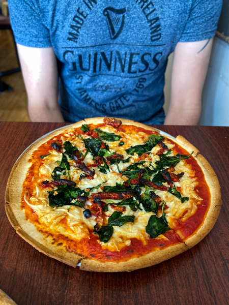 Tom, a white man in blue Guinness shirt, sitting in front of gluten free AsheVegan pizza with basil, sun dried tomatoes, sauce, vegan mozzarella from Manicomio Pizza Place in Asheville, NC