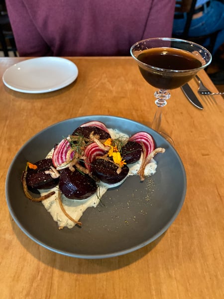 Plant Vegan Restaurant Asheville NC Beets on blue gray plate with fried onions on top and white sauce on bottom next to brown Manhattan cocktail