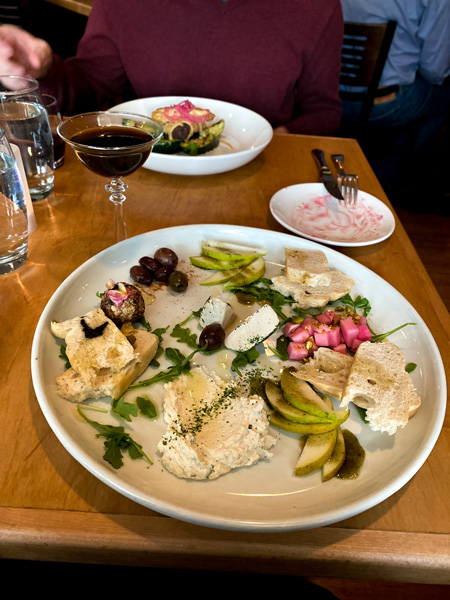 Plant Vegan Restaurant North Asheville Cheese Plate with white plate filled with vegan cheeses made from nuts paired with pears, pickled veggies like beets, hard bread, and olives on table with second entree blurred in background