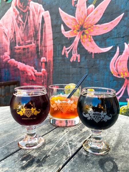 Wicked Weed Funkatorium with two sour beers and a cocktail on table in front of mural of man with pink flowers