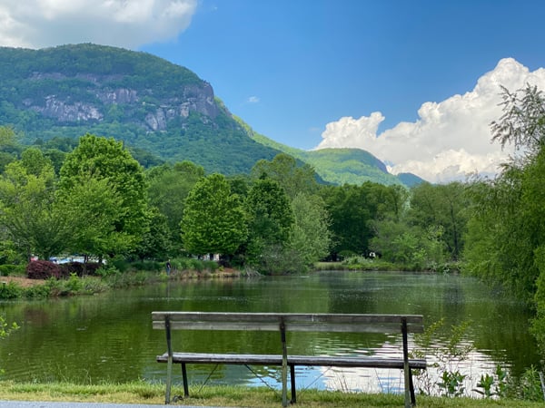 Lake Lure overlooking Chimney Rock in Western North Carolina with bench at lake, green trees, and blue cloudy sky