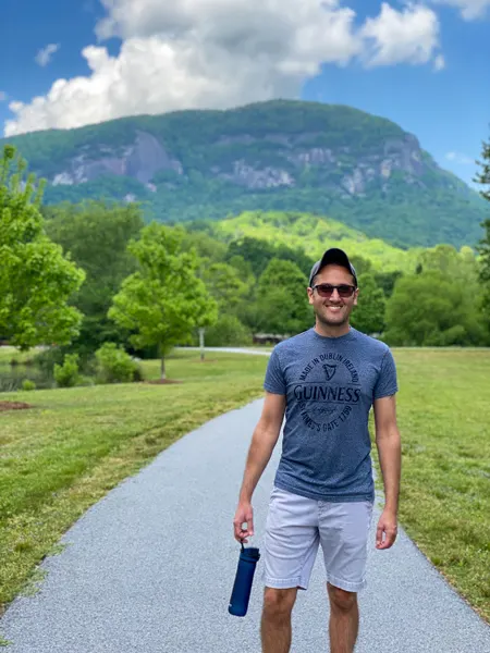 Lake Lure Trail with white brunette male in hat and blue short with cargo pants holding a water bottle on paved trail with Chimney Rock in background