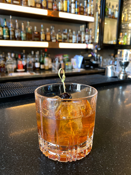 Times Bar and Coffee Shop Asheville Cocktail with brown-orange drink with cherry garnish on brown bar with liquors in background