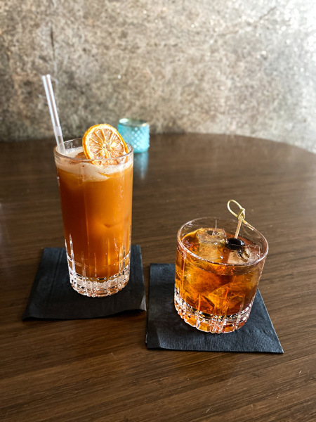 Times Bar Mocktail and cocktail in Asheville NC with two cocktails on brown table that are brownish in color, one in a low ball glass garnished with a black cherry and the other in a high ball glass garnished with lemon and straw