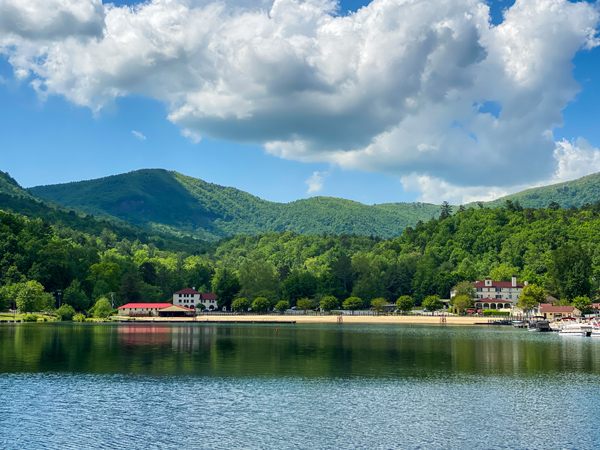 Lake Lure Beach NC with blue green lake water, sandy tan shore, blue and green mountains with trees, and blue cloudy sky