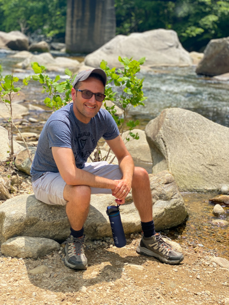 Rocky Broad River Walk in Chimney Rock NC with white brunette male in tan shorts and blue shirt with hat and sunglasses sitting on rocks by river