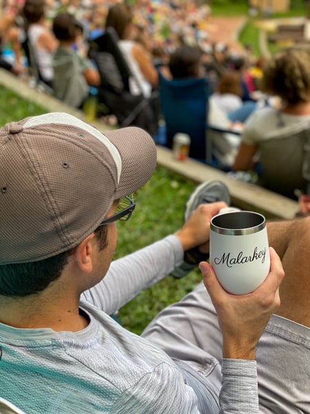 Shakespeare in the Park Asheville seating with white man in law chair wearing gray hat and holding Malarkey mug in front of Shakespeare stage