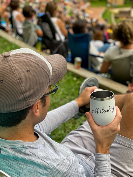 Tom, a white man in law chair wearing gray hat and holding Malarkey mug in front of stage for Shakespeare in the Park in Asheville, NC