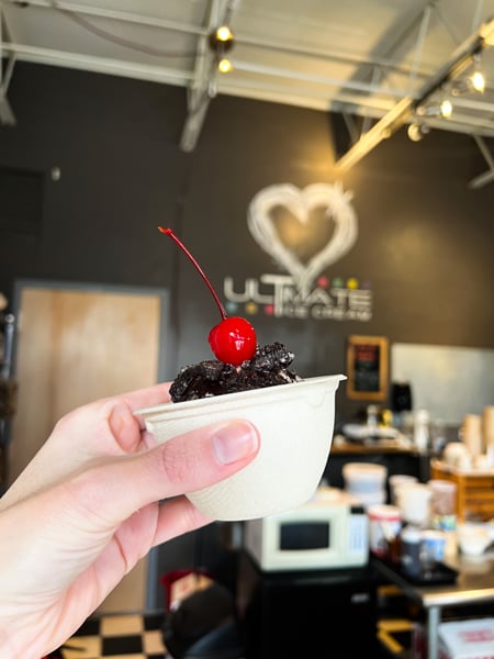 Ultimate Ice Cream Asheville North Carolina with white hand holding up kids' cup size of almond milk vanilla ice cream with chocolate Oreos and cherry on top in front of business heart logo