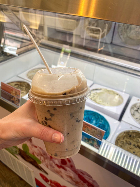 Village Scoop Chimney Rock NC with white hand holding dairy-free milkshake in front of ice cream counter filled with tubs of ice cream