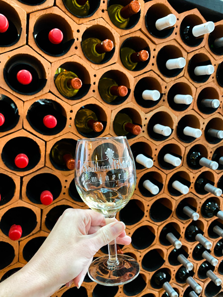 Southern Charm Winery Lincolnton NC with white hand holding up white wine glass in front of wine rank filled with bottles