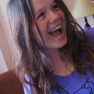 Dagney McKinney portrait of white light brunette with long hair and is laughing with an open mouth and wearing a purple t-shirt with lamp in background