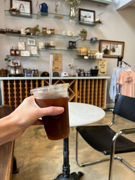 ShareWell Coffee Co Roastery in Flat Rock NC with white hand holding up iced coffee in to-go cup in front of merchandise like t shirts and mugs