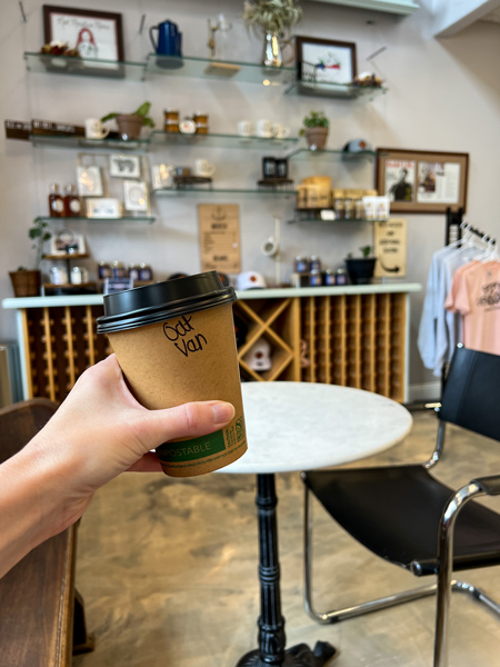 ShareWell Coffee Co Roastery near Hendersonville NC with white hand holding up oat milk latte in to-go cup in front of merchandise like t shirts and mugs