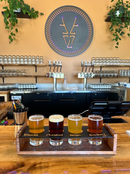 7 Clans Brewing Asheville NC with logo on light peach/orange wall, beer taps, and flight of 4 beers ranging from yellow to orange and brown in color