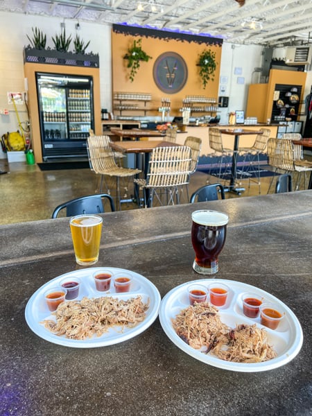 Garage BBQ 7 Clans Brewing Asheville with two styrofoam plates of pulled pork and pulled chicken with 4 BBQ sauces on each plate with two glasses of beer and brewery taproom in background