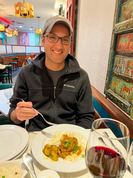 Mehfil Indian Restaurant Asheville NC with white male in hat and black fleece eating plate of spicy Indian food with glass of red wine