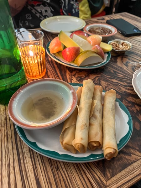Fried lumpia rolls on plate with opaque brown sauce and plate of apples and pears in background with candle on table at Neng Jr.'s Restaurant in West Asheville, NC