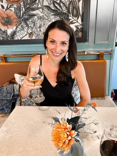 New Gemelli Restaurant Asheville NC with white brunette woman in a black dress holding a white wine glass with vase of flowers in front