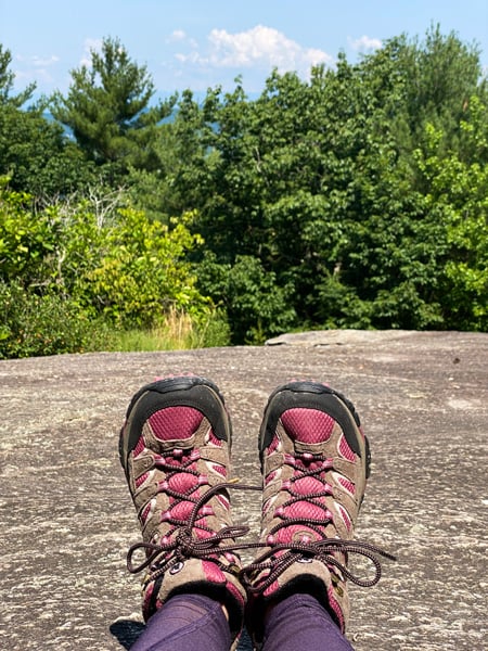 Glassy Mountain Trail Flat Rock NC with pair of legs in purple hiking leggings and fuchsia hiking shoes on flat rock at summit with views of clouds and green trees