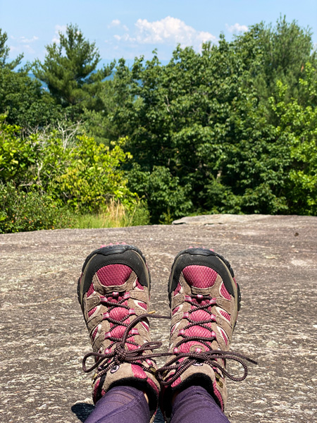 Glassy Mountain Trail Flat Rock NC with pair of legs in purple hiking leggings and fuchsia hiking shoes on flat rock at summit with views of clouds and green trees 