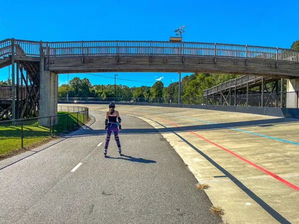 Mellowdrome Rollerblading Asheville NC with white brunette female in bike helmet on Carrier Park Velodrome which is a paved track with blue sky, green trees, and wooden bridge to get there