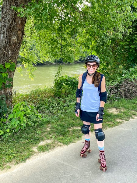Christine, a white brunette female in maroon squad skates with helmet, pads, sunglasses, and workout top and skirt by French Broad River on the RAD Riverwalk roller skating in Asheville, NC 
