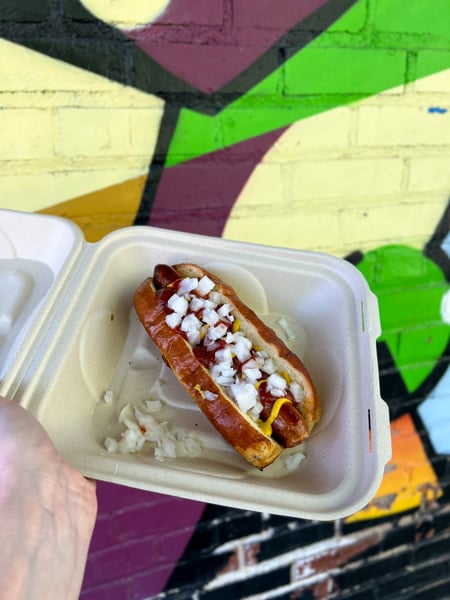 Chop Shop Food Truck in Asheville, North Carolina with white hand holding up a takeaway container with a hot dog with mustard and onions on top