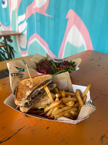 Foothills Asheville Food Truck At Hi Wire Beer Garden NC with sandwich filled with smashed burger, egg, shredded meat and side of fries will pulled pork salad in background