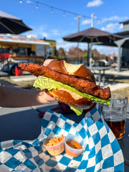 Haus Heidelberg Food Truck Near Asheville NC with white hand holding up schnitzel on pretzel bread at picnic table with food truck in background