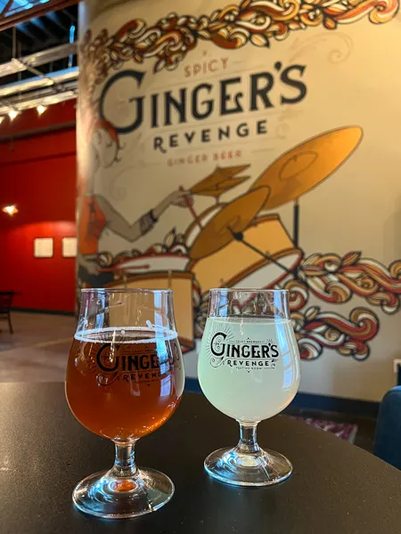 Gingers Revenge in Asheville, NC with ginger beer on brown table. There are two glasses where one has dark brown ginger beer and one has light yellow ginger beer with a mural of red haired drummer in the background