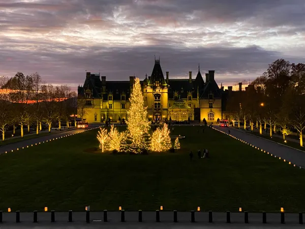 Biltmore House at night for Christmas with Blue Ridge Mountain sunset sky with purple clouds and yellow lighted trees out front in Asheville NC