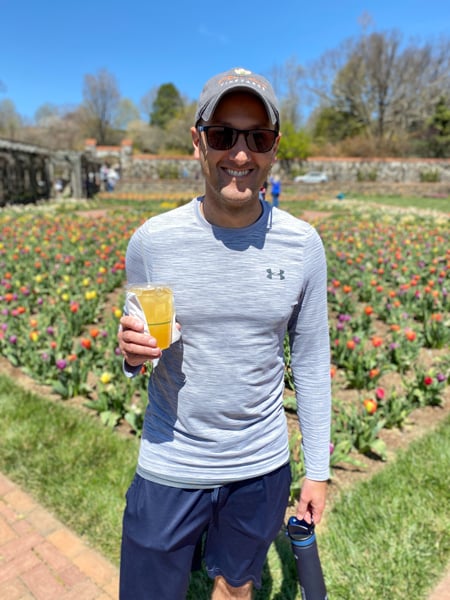 The Conservatory Cafe At Biltmore in Asheville NC with white brunette male wearing hat, gray top, blue shorts, and holding mimosa like drink in plastic cup in gardens with blue sky