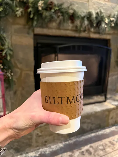 The Kitchen Cafe at the Village Hotel at Biltmore Coffee in to-go held up by white woman's hand in front of gas fireplace with holiday decor