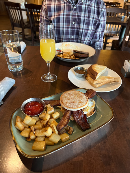 Village Social Biltmore Breakfast in Asheville, NC with blue gray plate filled with egg, bacon, english muffin breakfast sandwich, roasted potatoes, and ketchup with mimosa and gluten-free toast on white plate in background