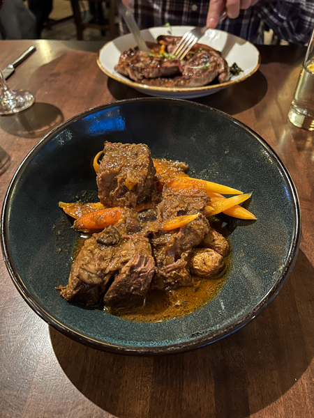 Village Social Biltmore Estate Restaurant with Pot Roast in black bowl with carrots, gravy, and potatoes with steak dish blurred in background