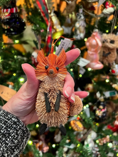 A white hand holding up a fox ornament in front of decorated Christmas tree at Winter Lights at The NC Arboretum in Asheville, NC