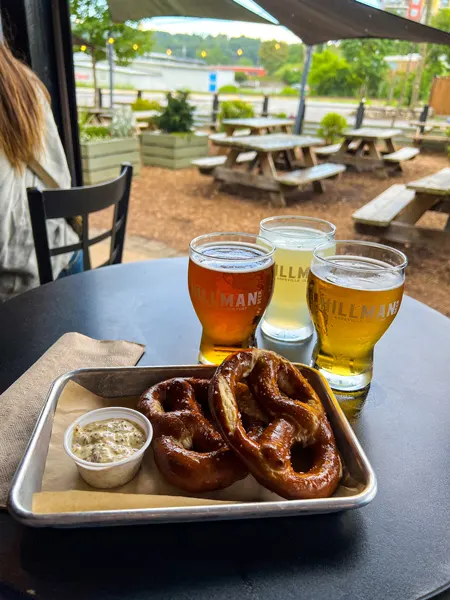Hillman Beer in Asheville's Biltmore Village food tray with two brown pretzels and dip along with cider and amber beer on black table top under covered seating that is half outside with blurred picnic tables in the background