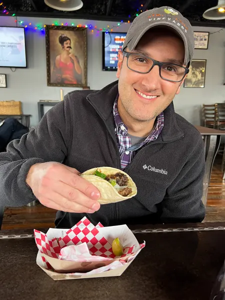 The Grey Eagle Taqueria at Big Pillow Brewing in Hot Springs North Carolina with white brunette male wearing a hat, black fleece, and glasses holding up a corn tortilla taco