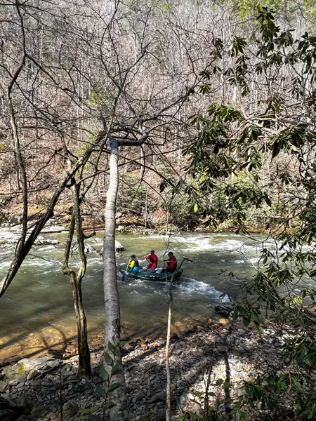 Whitewater rafting in Hot Springs, NC with image of three people in raft going over small rapids along a river with trees around it