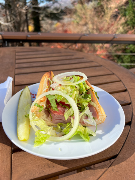 Apollo Flame takeout in Asheville with image of open ham and Italian meats sub with large onion, lettuce, tomato, and pickle on side on white plate on outdoor table