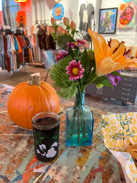 Great Wild Nowhere Art Bar with orange pumpkin, cup of red wine, and blue flower vase with flowers on colorful picnic table with retail clothing store in the background