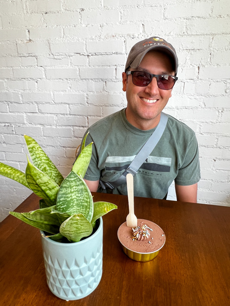 Ivy and the Poet Cafe in Marshall NC vegan and gluten-free chocolate cake dessert on table with green plant and white brunette male wearing green t-shirt, hat and sunglasses