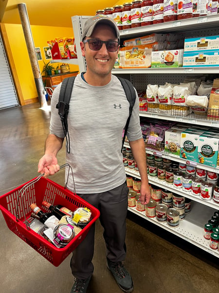 Madison Natural Foods Store in Marshall North Carolina with white brunette male in sunglasses and hiking shorts and top with hat carrying red grocery basket filled with wine, cheese, and olives