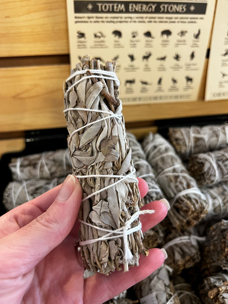 Of Wand & Earth Shop in Marshall North Carolina with white hand holding up wrapped bundle of greenish sage in front of display